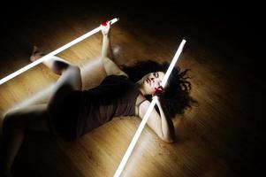 Curly brunette plus size model with two long led lamp at black room lying on flat. photo