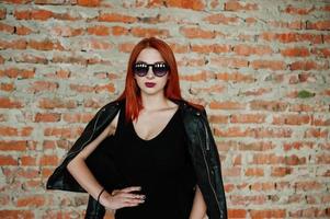 chica elegante de pelo rojo con gafas de sol vestidas de negro, contra un lugar abandonado con paredes de ladrillo. foto