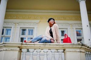 Red haired girl in glasses and hat with red handbag posed near vintage house. photo