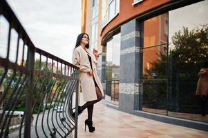 mujer sexy de pelo negro con gafas y abrigo posado contra un edificio con ventanas modernas. foto