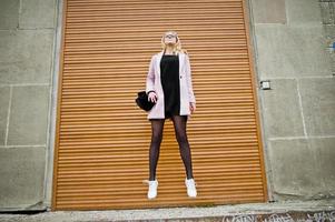 Blonde girl at glasses and pink coat with handbag jump against shutters. photo