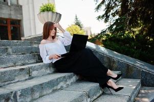 Good-looking young woman in white blouse, wide black pants and black classic high heels sitting on stairs and working on her laptop. photo