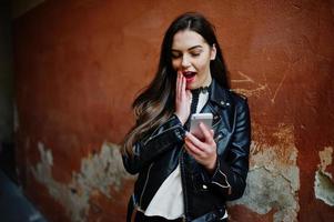 elegante chica morena vestida con chaqueta de cuero y pantalones cortos con teléfono móvil en las manos contra la pared naranja. foto