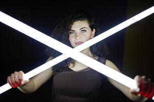 Curly brunette plus size model with two long led lamp at black room. photo