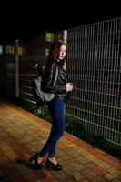 retrato nocturno de una modelo de niña vestida con jeans y chaqueta de cuero contra una verja de hierro. foto