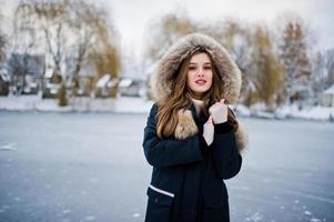 Beautiful brunette girl in winter warm clothing. Model on winter jacket against frozen lake at park. photo