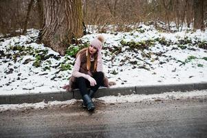 Stylish girl in fur coat and headwear at winter day on sitting border ofroad. photo