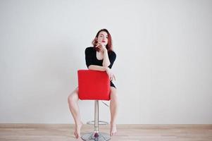 Red haired girl on black dress tunic sitting on red chair against white wall at empty room. photo