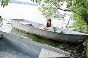 retrato de una mujer atractiva con vestido de lunares negros, chal blanco y gafas leyendo un libro en un bote en un lago. foto