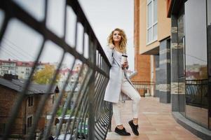 Stylish curly blonde model girl wear on white with cup of coffee at hand posing against railings. photo