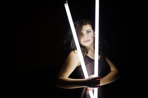 Curly brunette plus size model with two long led lamp at black room. photo