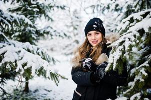 Beautiful brunette girl in winter warm clothing. Model on winter jacket and hat near pine trees.