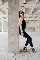 Red haired stylish girl in sunglasses wear in black, against abadoned place. photo