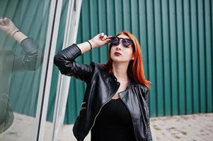 chica elegante de pelo rojo con gafas de sol en negro, contra grandes ventanales. retrato de moda callejera. foto