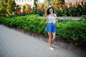 ropa de niña con estilo rizado en falda de jeans azules, blusa y gafas posadas en la calle de la ciudad con una taza de café. foto