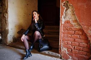 Stylish brunette girl wear on leather jacket and shorts with backpack sitting on old stairs against orange wall. photo