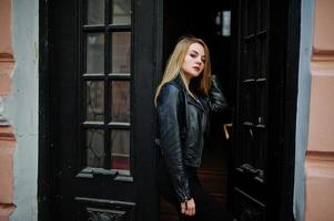 Elegant blonde girl wear on black leather jacket posing at streets of town background old doors. photo