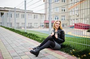 Blonde fashionable girl in long black leather coat against iron fence at street. photo