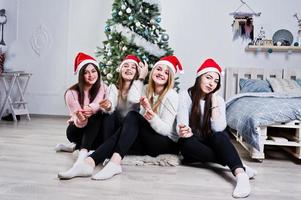 Four cute friends girls wear on warm sweaters,  black pants and red santa hats against new year tree with christmas decoration at white room and hold bengal lights at hands. photo