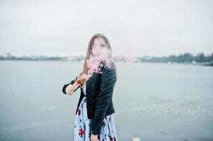 chica elegante con chaqueta de cuero sostiene una bengala de humo rosa en el día de invierno contra el lago congelado. foto