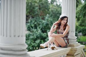 Increíbles piernas largas con tacones altos, ropa de niña con sombrero sentado cerca de columnas de edificios antiguos. foto