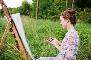 Portrait of a gorgeous happy young woman in beautiful dress sitting on the grass and painting on paper with watercolors. photo