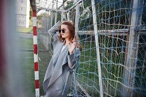 Girl in gray coat with sunglasses at small street stadium football gates. photo