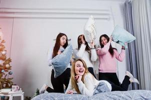 Four cute friends girls wear on warm sweaters and black pants on bed at new year decorated room on studio, play with pillows. photo