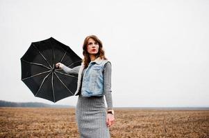 Portrait of brunette curly girl in jeans jacket with black umbrella at field. photo