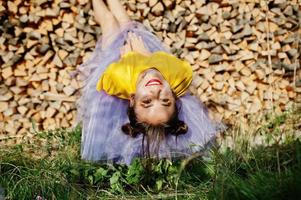 Young funny girl with bright make-up, like fairytale princess, wear on yellow shirt and violet skirt lying against wooden background. photo