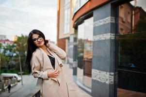 mujer sexy de pelo negro con gafas y abrigo posado contra un edificio con ventanas modernas. foto