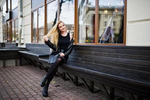 Blonde fashionable girl in long black leather coat sitting on bench. photo