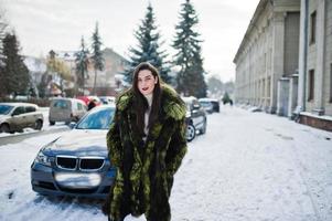 Brunette girl in green fur coat at street of city background car in winter day. photo