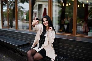 mujer sexy de pelo negro con gafas y abrigo sentado en un banco con teléfono móvil a mano y haciendo selfie. foto