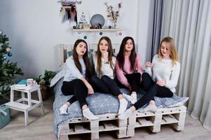 Four cute friends girls wear on warm sweaters and black pants on bed at new year decorated room on studio. photo