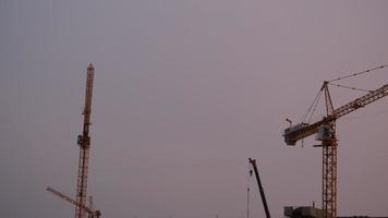tornkranar som arbetar på en byggarbetsplats lyfter en last vid höghus i skymningen. en stor byggarbetsplats med upptagna kranar. video