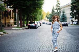 Portrait of a beautiful model in striped overall posing with hat and a backpack on a street with trees in a town. photo