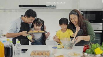 Happy family mom dad and kids siblings cooking together, parents teaching children son daughter cooking fresh vegetable salad and croissant prepare healthy food in modern kitchen interior together video