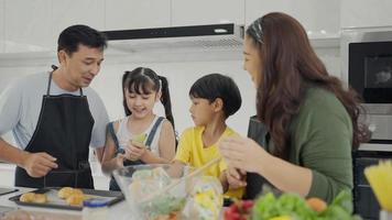 feliz familia mamá papá y niños hermanos cocinando juntos, padres enseñando a los niños hijo hija cocinando ensalada de verduras frescas y croissant preparar comida saludable en el interior de la cocina moderna juntos video