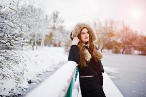 Beautiful brunette girl in winter warm clothing. Model on winter jacket against frozen lake at park. photo