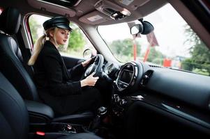 Portrait of beautiful blonde sexy fashion woman model in cap and in all black with bright makeup sit and drive red city car. photo