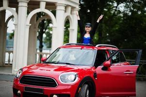 Portrait of beautiful blonde sexy fashion woman model in cap and in all black with bright makeup in red city car open sunroof. photo