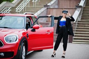 Portrait of beautiful blonde sexy fashion woman model in cap and in all black, blue corset, with bright makeup near red city car. photo