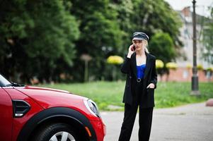Portrait of beautiful blonde sexy fashion woman model in cap and in all black with bright makeup near red city car, with mobile phone. photo