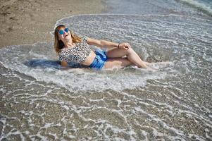 Beautiful model relaxing on a beach of sea, wearing on jeans short, leopard shirt and sunglasses. photo