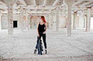 chica elegante de pelo rojo con gafas de sol vestidas de negro, contra un lugar abandonado. foto