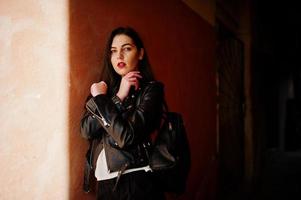 Stylish brunette girl wear on leather jacket and shorts with backpack against orange wall at shadows. photo