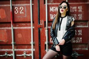 Stylish brunette girl wear on leather jacket and shorts, sunglasses against shipping container. photo