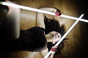 Curly brunette plus size model with two long led lamp at black room lying on flat. photo