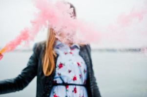 Stylish girl in leather jacket hold pink smoke flare at winter day against frozen lake. photo